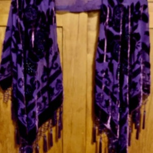 Peut inclure: Un châle en velours violet avec un motif floral et des franges perlées. Le châle est drapé sur une surface en bois.