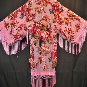 May include: A long, light pink kimono-style robe featuring a dense, dark red and green floral pattern. The garment has wide, open sleeves and a straight hem, both finished with a wide pink velvet trim and long, dangling pink fringe detailing.