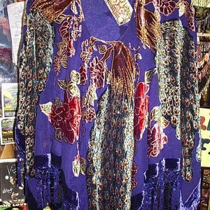 Peut inclure: Une tunique violette à manches longues avec un col en V, ornée d'un motif de paon et de fleurs. La tunique a des accents de velours et des franges à l'ourlet. Le motif incorpore des couleurs rouge, or et sarcelle.