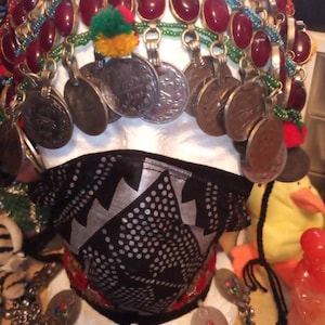 Puede incluir: Un tocado de cabeza de cuentas plateadas y rojas con monedas colgantes y piedras rojas. El tocado se lleva sobre una cabeza de maniquí blanca con una máscara facial de patrón blanco y negro.