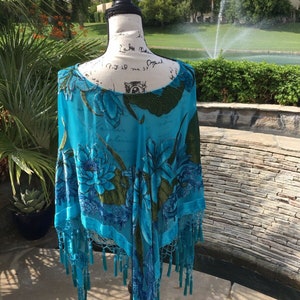 Peut inclure: Un poncho bleu turquoise avec un motif floral et une bordure à franges. Le tissu transparent présente de grandes fleurs bleues avec des feuilles vertes et est présenté sur un mannequin. Le poncho est un accessoire élégant.