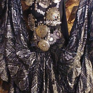 Puede incluir: Una falda negra y plateada con un patrón geométrico. La falda se combina con un conjunto de joyería de fantasía plateada y negra, que incluye un collar, un cinturón y una pieza de pecho.