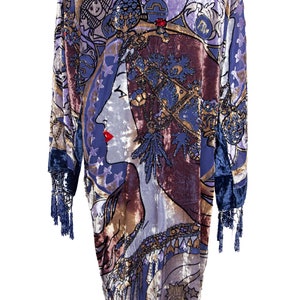 Peut inclure: Une robe en velours bleu avec un imprimé coloré d'un visage de femme et de symboles astrologiques. La robe a une bordure en frange et est exposée sur un mannequin.