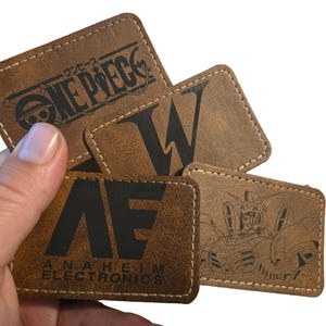 May include: A collection of brown leather patches with various designs. One patch features the text "ONE PIECE" in black, another displays a large black "W", and a third reads "AE ANAHEIM ELECTRONICS". The fourth patch has a robot illustration.