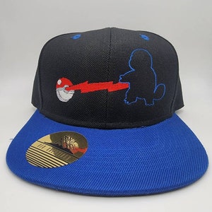 May include: Black and blue baseball cap with an embroidered image of a Pokemon character, Snorlax, with a Pokeball and red lightning bolt. The cap has a gold sticker on the underside of the brim.