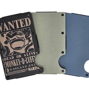 May include: Three metal wallets in black, green, and blue. The black wallet features a "WANTED" poster design with the text "MONKEY D. LUFFY" and a bounty of 3,000,000,000. The wallets have a minimalist design with visible screws.