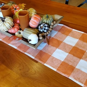 May include: A wooden table is adorned with a fall-themed centerpiece. A wooden tray holds decorative pumpkins, gourds, and fall foliage. An orange and white checkered table runner adds a seasonal touch to the table setting.