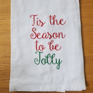 May include: White kitchen towel with red and green embroidered text that reads "Tis the Season to be Jolly".