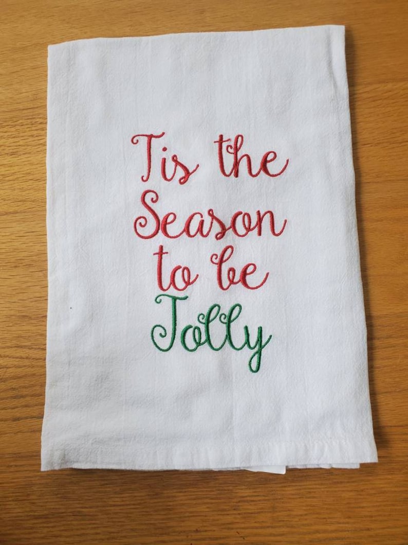 May include: White kitchen towel with red and green embroidered text that reads "Tis the Season to be Jolly".