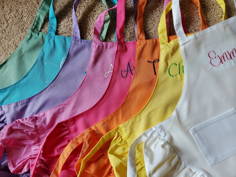 May include: A set of ten colorful aprons with ruffled edges. Each apron has a different color and is personalized with a name in white or green embroidery. The aprons are arranged in a rainbow pattern.