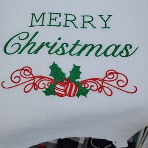 May include: White kitchen towel with green embroidered text "Merry Christmas" and a red and white striped ornament with green holly leaves.