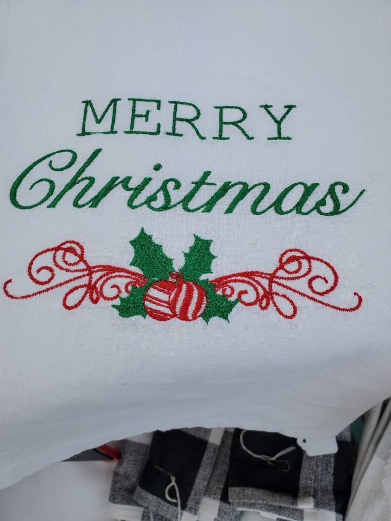 May include: White kitchen towel with green embroidered text "Merry Christmas" and a red and white striped ornament with green holly leaves.