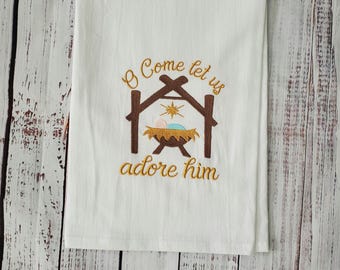 May include: White kitchen towel with embroidered text and a nativity scene. The text reads "O Come let us adore him" in gold thread. The nativity scene features a brown stable and a baby in a manger.