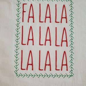 May include: A white fabric with red embroidery of the phrase "Fa La La La La La La" in a green embroidered frame.