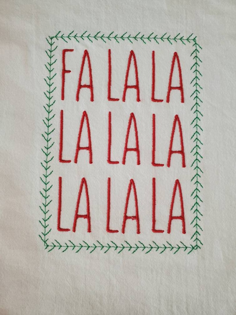 May include: A white fabric with red embroidery of the phrase "Fa La La La La La La" in a green embroidered frame.