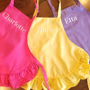 May include: Three ruffled aprons in pink, yellow, and purple with white embroidered names: Charlotte, Halle, and Etta.