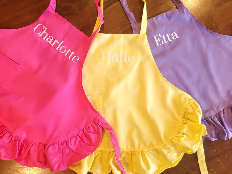 May include: Three ruffled aprons in pink, yellow, and purple with white embroidered names: Charlotte, Halle, and Etta.