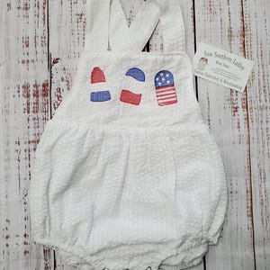 May include: White seersucker baby romper with embroidered red, white, and blue popsicle designs. Features adjustable straps and snap closures at the bottom. The romper is designed for comfort and style.