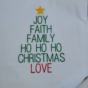May include: White kitchen towel with green embroidered text in the shape of a Christmas tree. The text reads "Joy, Faith, Family, Ho Ho Ho, Christmas, Love". The top of the tree is a yellow star.