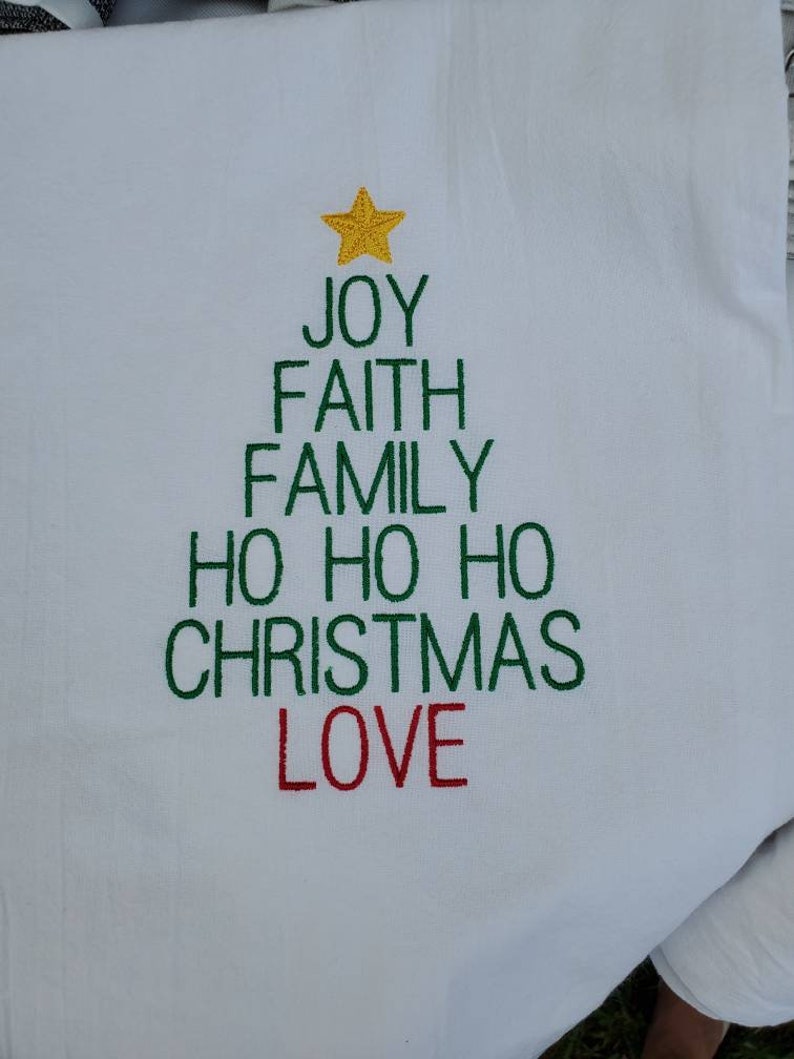 May include: White kitchen towel with green embroidered text in the shape of a Christmas tree. The text reads "Joy, Faith, Family, Ho Ho Ho, Christmas, Love". The top of the tree is a yellow star.