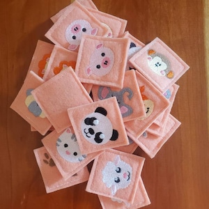 May include: A set of felt squares with embroidered animal faces. The squares are peach colored and feature a variety of animals including a pig, panda, cat, sheep, and elephant.