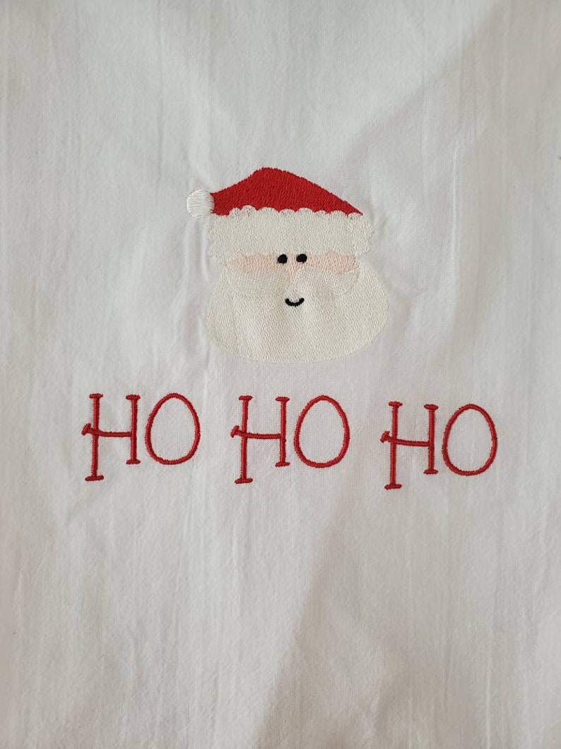 May include: White fabric with a red embroidered Santa Claus face and the text "HO HO HO" in red.
