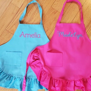 May include: Two personalized children's aprons with ruffled edges. One apron is turquoise blue with the name "Amelia" embroidered in pink. The other apron is hot pink with the name "Madelyn" embroidered in blue.