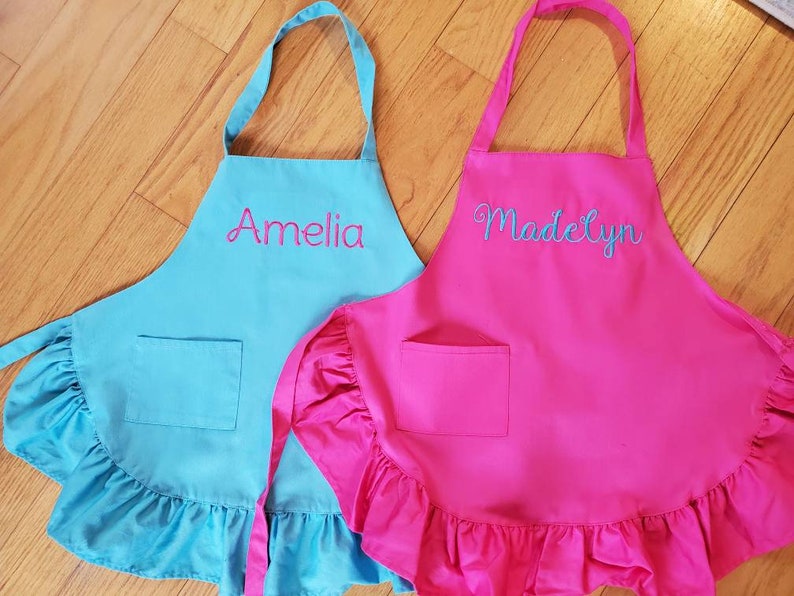 May include: Two personalized children's aprons with ruffled edges. One apron is turquoise blue with the name "Amelia" embroidered in pink. The other apron is hot pink with the name "Madelyn" embroidered in blue.