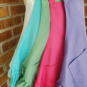 May include: Four ruffled aprons in different colors: white, turquoise, green, and purple. Each apron has a single pocket and adjustable straps.