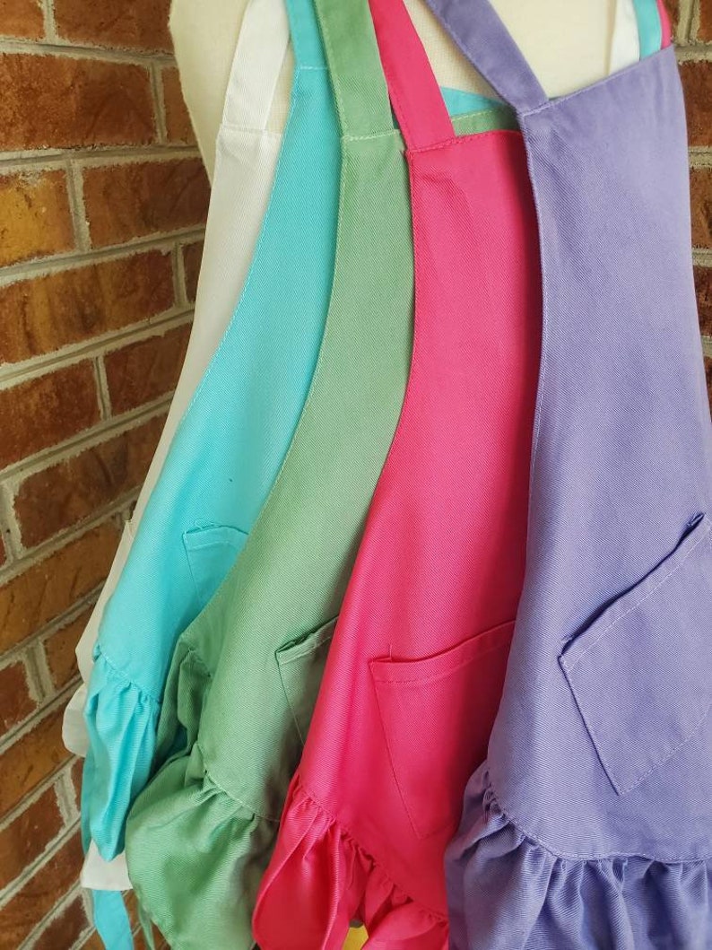 May include: Four ruffled aprons in different colors: white, turquoise, green, and purple. Each apron has a single pocket and adjustable straps.