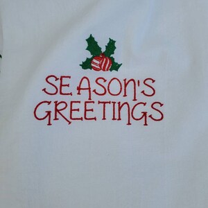 May include: White fabric with red embroidered text that says "Season's Greetings" with a green holly sprig and red and white striped ornaments above the text.