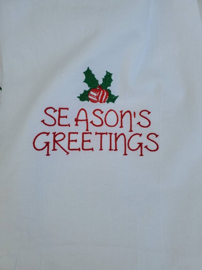 May include: White fabric with red embroidered text that says "Season's Greetings" with a green holly sprig and red and white striped ornaments above the text.