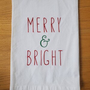 May include: White kitchen towel with red embroidery that reads "Merry & Bright" with a green ampersand.