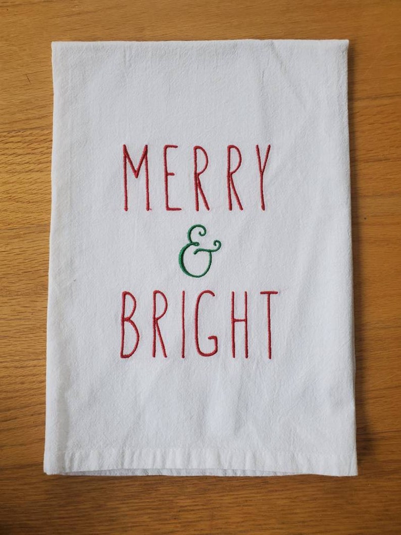 May include: White kitchen towel with red embroidery that reads "Merry & Bright" with a green ampersand.