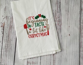 May include: White kitchen towel with red embroidered text that reads "It's beginning to taste a lot like Christmas!" with a red holly sprig, a red spatula, and a silver whisk.