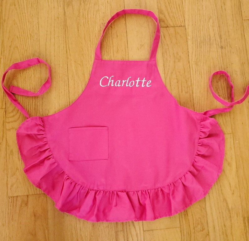 May include: Pink ruffled apron with a pocket and the name "Charlotte" embroidered in white.