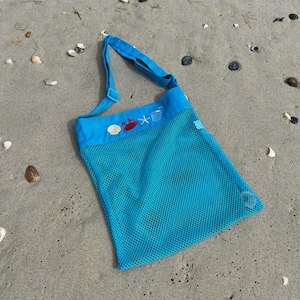 May include: A blue mesh beach bag with a white strap and embroidered images of a crab, starfish, and bucket. The bag is lying on a sandy beach.