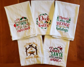 May include: Set of six white kitchen towels with embroidered Christmas-themed sayings. The sayings include "It's beginning to taste a lot like Christmas", "Deck the halls", "There's no place like home for the holidays", "O come let us adore him", and "Dear Santa just bring wine".