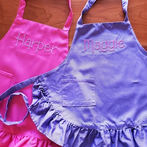 May include: Two personalized ruffled aprons, one pink with the name "Harper" embroidered on it and one purple with the name "Maggie" embroidered on it.