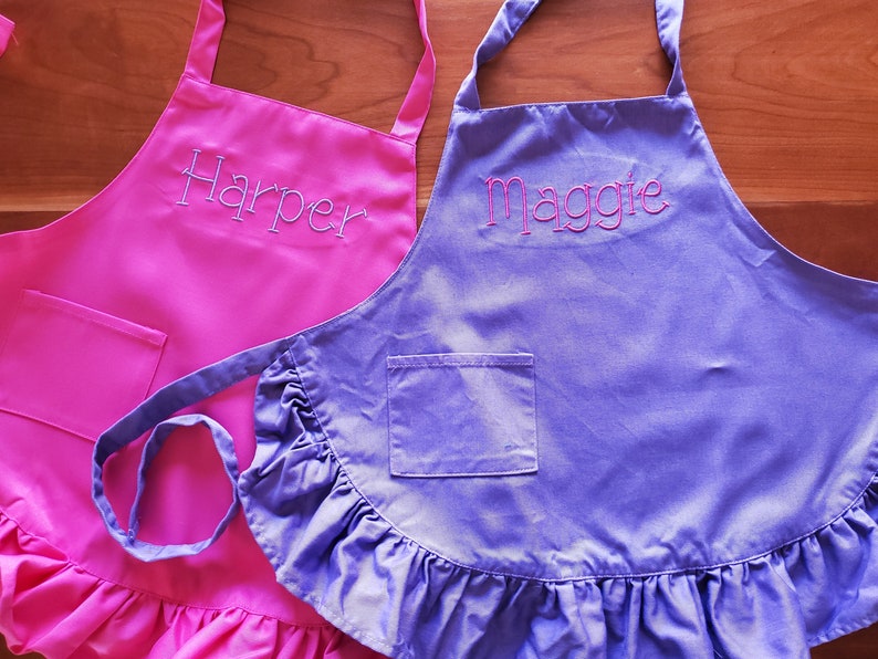 May include: Two personalized ruffled aprons, one pink with the name "Harper" embroidered on it and one purple with the name "Maggie" embroidered on it.