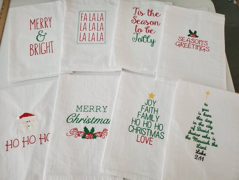 May include: Set of 10 white kitchen towels with red and green embroidered Christmas designs. The designs include "Merry & Bright", "Fa La La La La La", "Tis the Season to be Jolly", "Season's Greetings", "Ho Ho Ho", "Merry Christmas", "Joy Faith Family Ho Ho Ho Christmas Love", and a Christmas tree with the text "To you is born this day in the city of David a Savior who is the Messiah the Lord Luke 2:11".