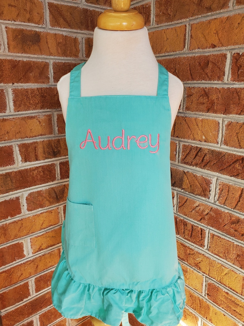 May include: A turquoise child's apron with a ruffled hem and a pocket. The apron has the name "Audrey" embroidered on the front in pink thread.