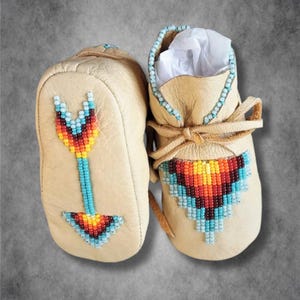 May include: A pair of beige leather baby moccasins with beaded arrow designs. The moccasins are decorated with turquoise, red, orange, and yellow beads.