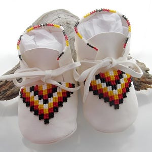 May include: White leather baby moccasins with beaded trim. The trim is a geometric pattern in red, yellow, black, and white.