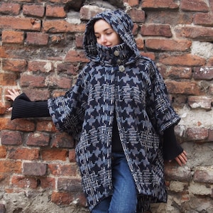 Peut inclure: Un manteau à capuche en tissu à motif pied-de-poule noir et blanc avec un col montant et une fermeture à boutons. Le manteau est porté par une personne debout contre un mur en briques.