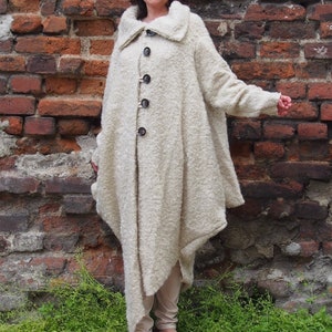 May include: A long, off-white, fuzzy coat with a large collar and button closure. The coat is asymmetrical and has a flowing, draped design. The model is wearing red shoes.