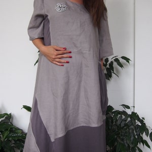 May include: A gray linen dress with a decorative flower brooch. The dress has a loose fit and a flowing skirt. The dress is asymmetrical and has a longer hem in the back.
