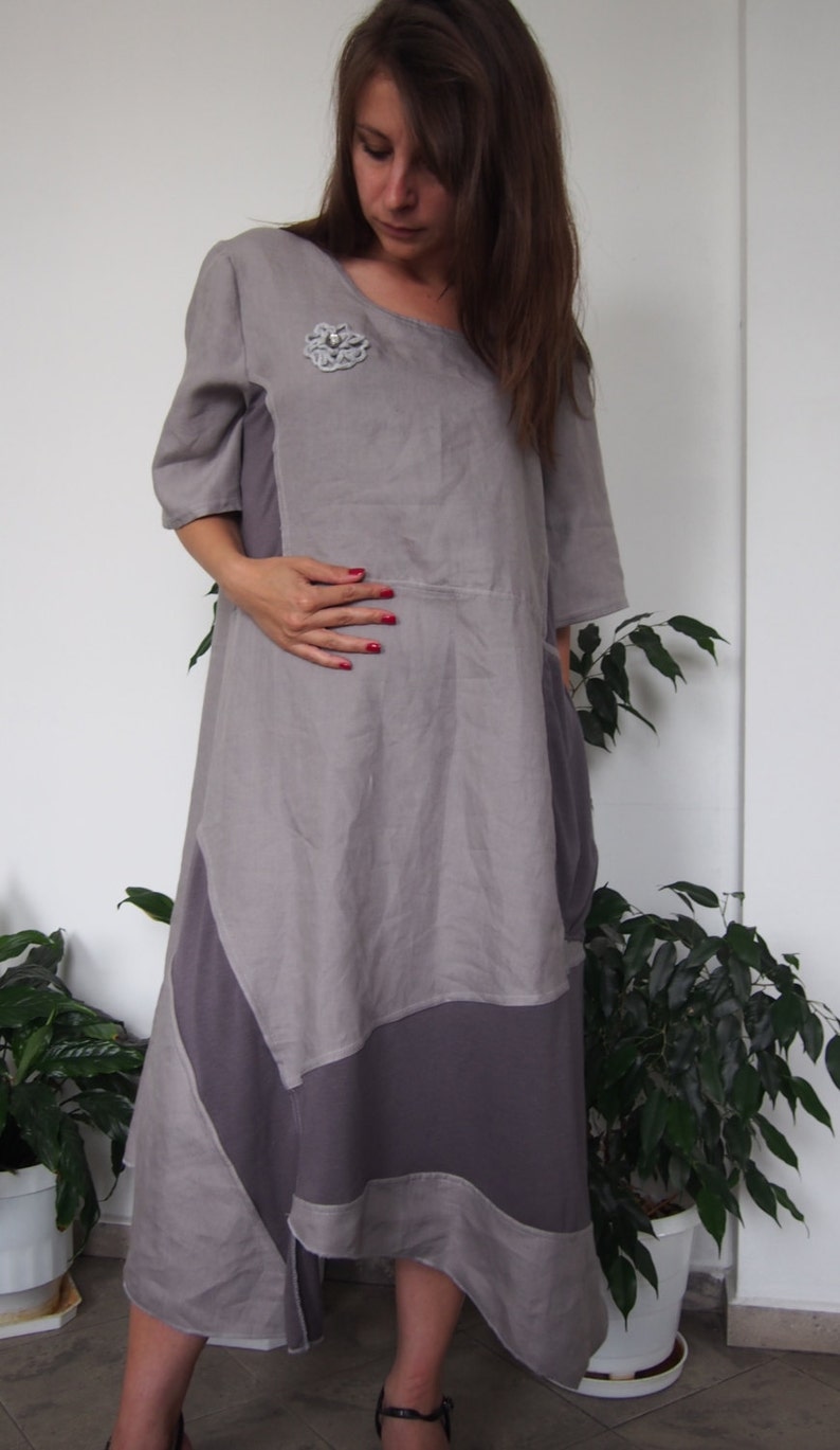 May include: A gray linen dress with a decorative flower brooch. The dress has a loose fit and a flowing skirt. The dress is asymmetrical and has a longer hem in the back.