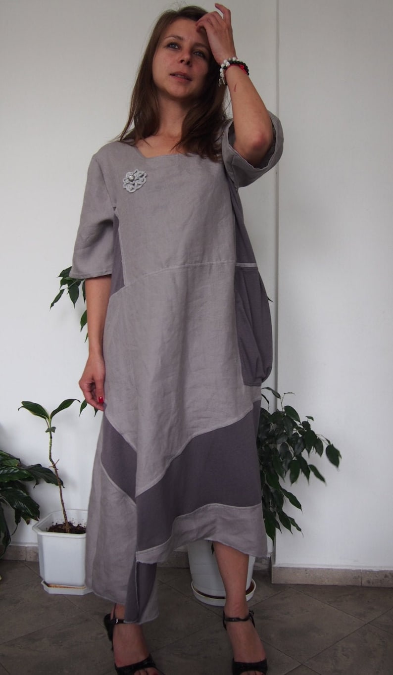 May include: A woman wearing a gray and purple asymmetrical dress with a flower brooch. She is wearing black sandals and has her hair pulled back.
