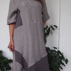 May include: A gray linen dress with a geometric design and a silver flower brooch. The dress has a loose fit and a flowing skirt.